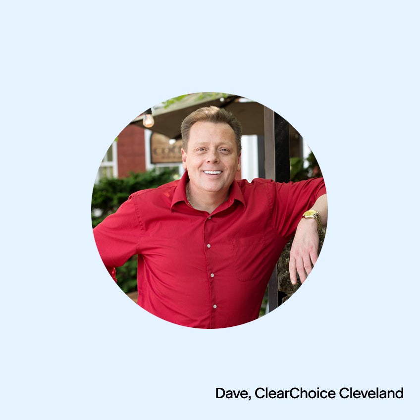 An image of a man in a red shirt smiling while leaning on a wall. 