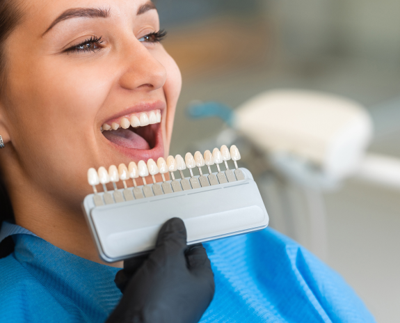 A patient sitting in a dentist's chair and smiling while the dentist compares a tooth color chart with her original tooth color.