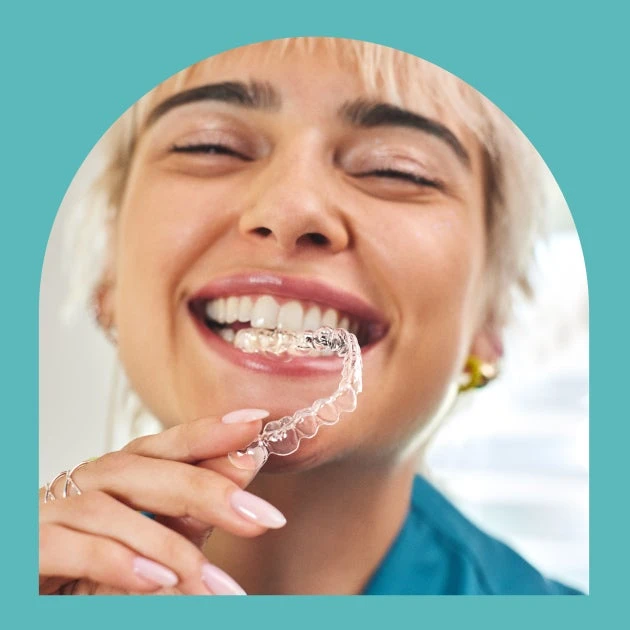 A woman with braces on her teeth is holding a clear retainer in her mouth.