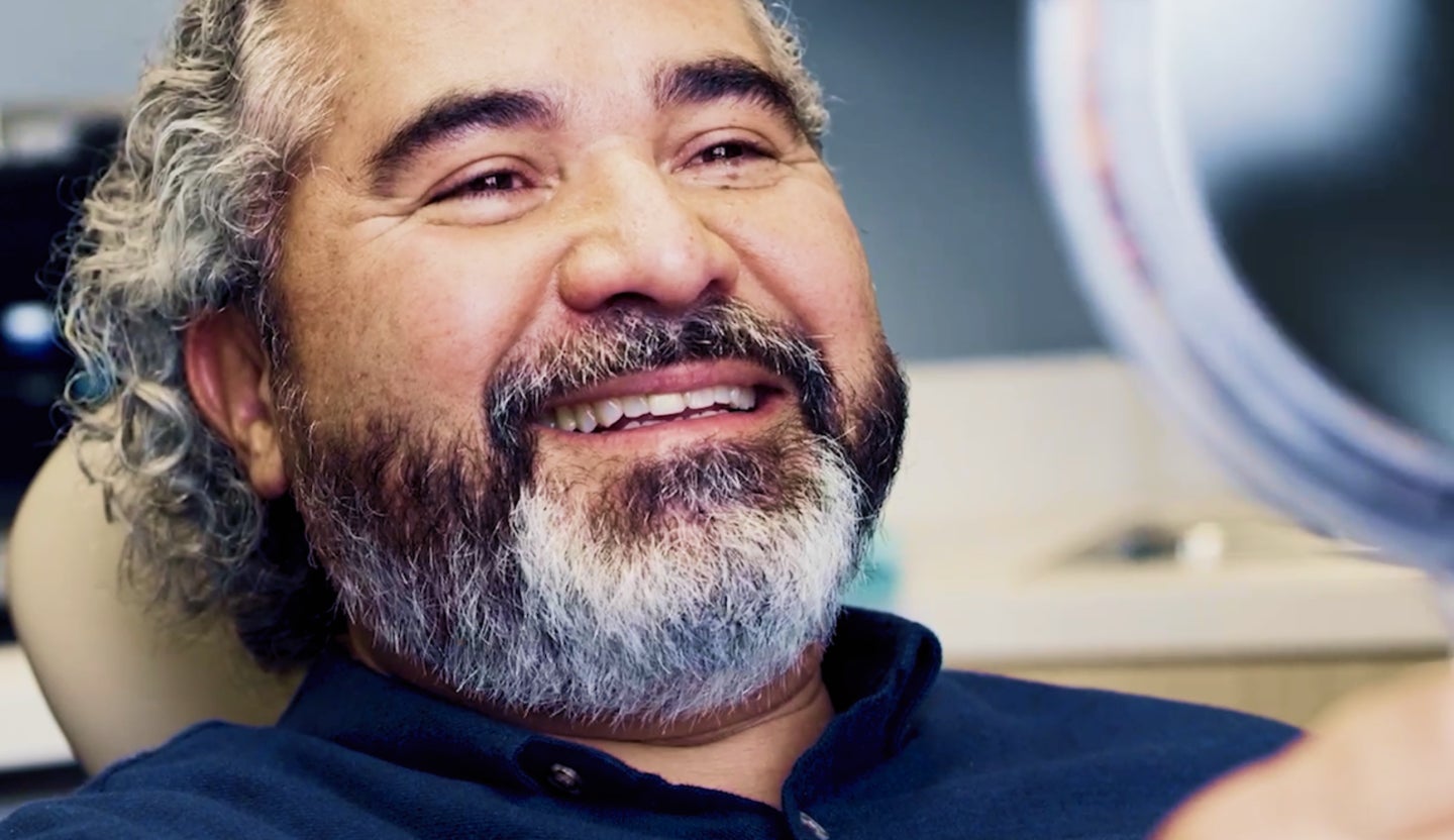 A cheerful Aspen Dental patient smiling while holding a mirror after a successful wisdom tooth extraction, seated comfortably in a modern dental clinic.