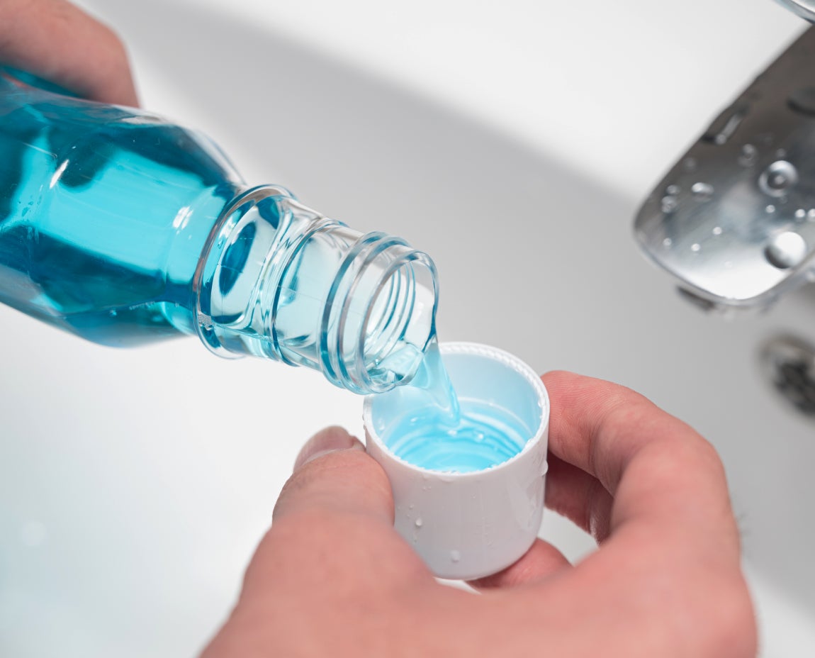 Close-up of a person pouring blue mouthwash into a cap over a bathroom sink, representing a step in maintaining oral hygiene. The vibrant blue color of the mouthwash adds a fresh, clean aesthetic, aligning with Aspen Dental’s emphasis on complete dental care.