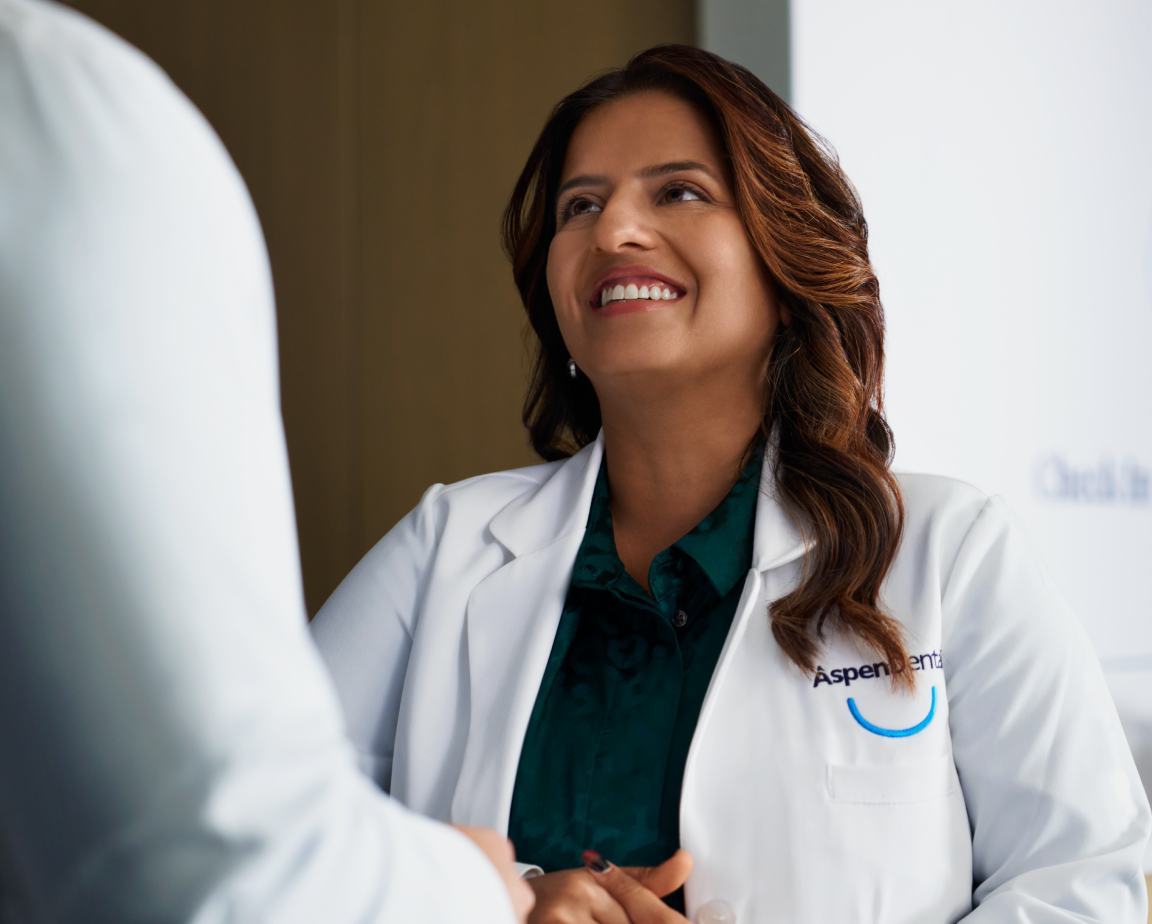 A smiling healthcare professional in a white coat with "Aspen Dental" logo visible on the pocket, stands and smiles at another person partially visible in the image.