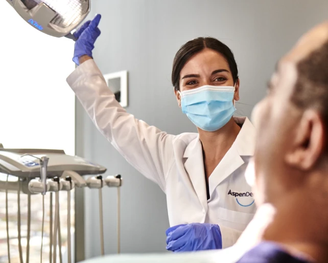 The Aspen Dental dentist wearing a mask and gloves holds up a dental light to examine a patient's teeth.