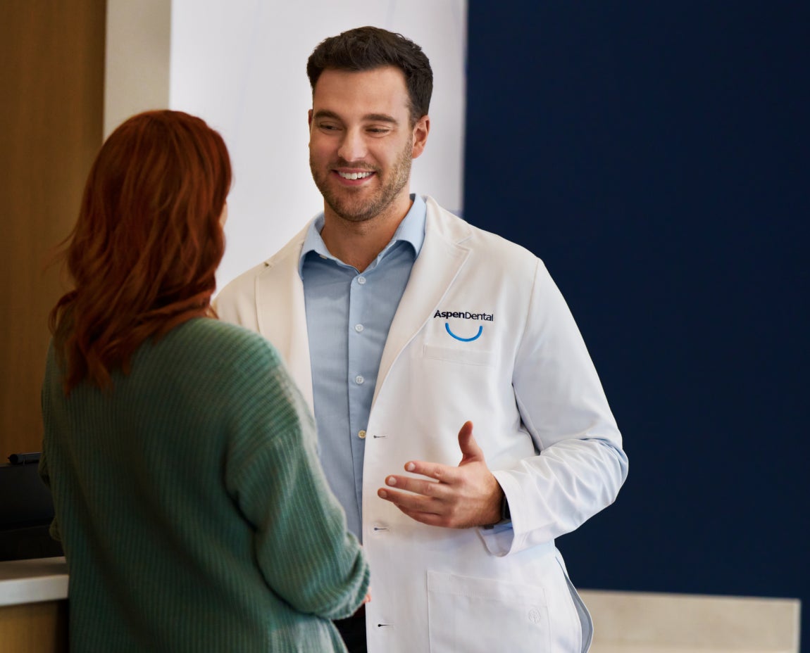 Aspen Dental dentist in a white coat smiling and consulting with a female patient wearing a green sweater in a modern dental office setting.
