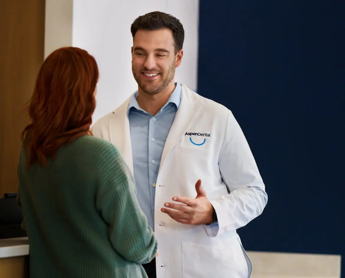 Aspen Dental dentist in a white coat smiling and consulting with a female patient wearing a green sweater in a modern dental office setting.