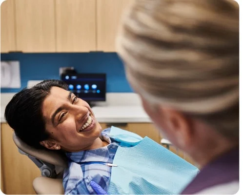 A person lies back in a dental chair smiling at another person in the foreground, who appears to be a dental professional in a clinic setting.