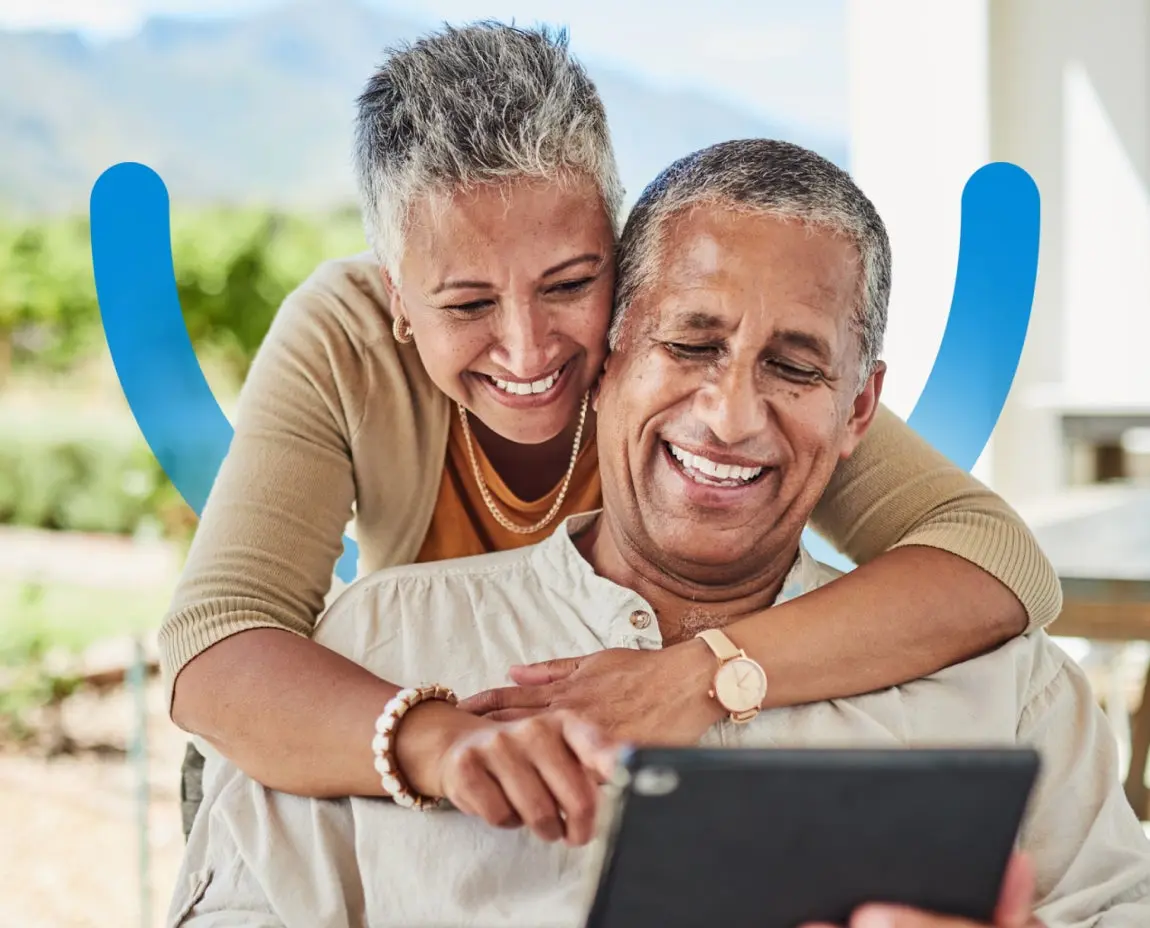 A woman is hugging a man while he looks at a tablet.
