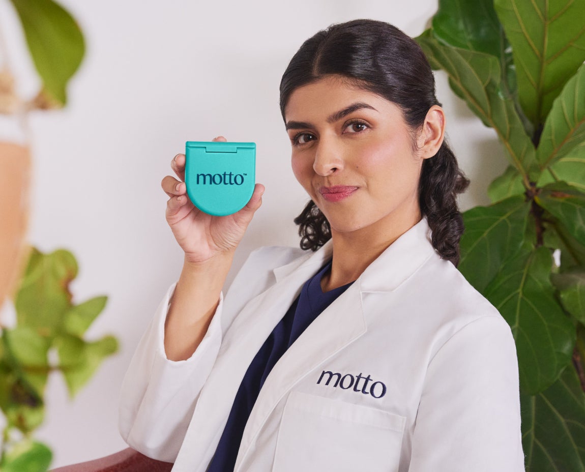 A dentist in a Motto branded white coat holding a teal container labeled "motto" against a backdrop of green plants.