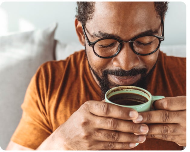 A person wearing glasses and an orange shirt holds a cup close to his face, smelling the beverage inside.