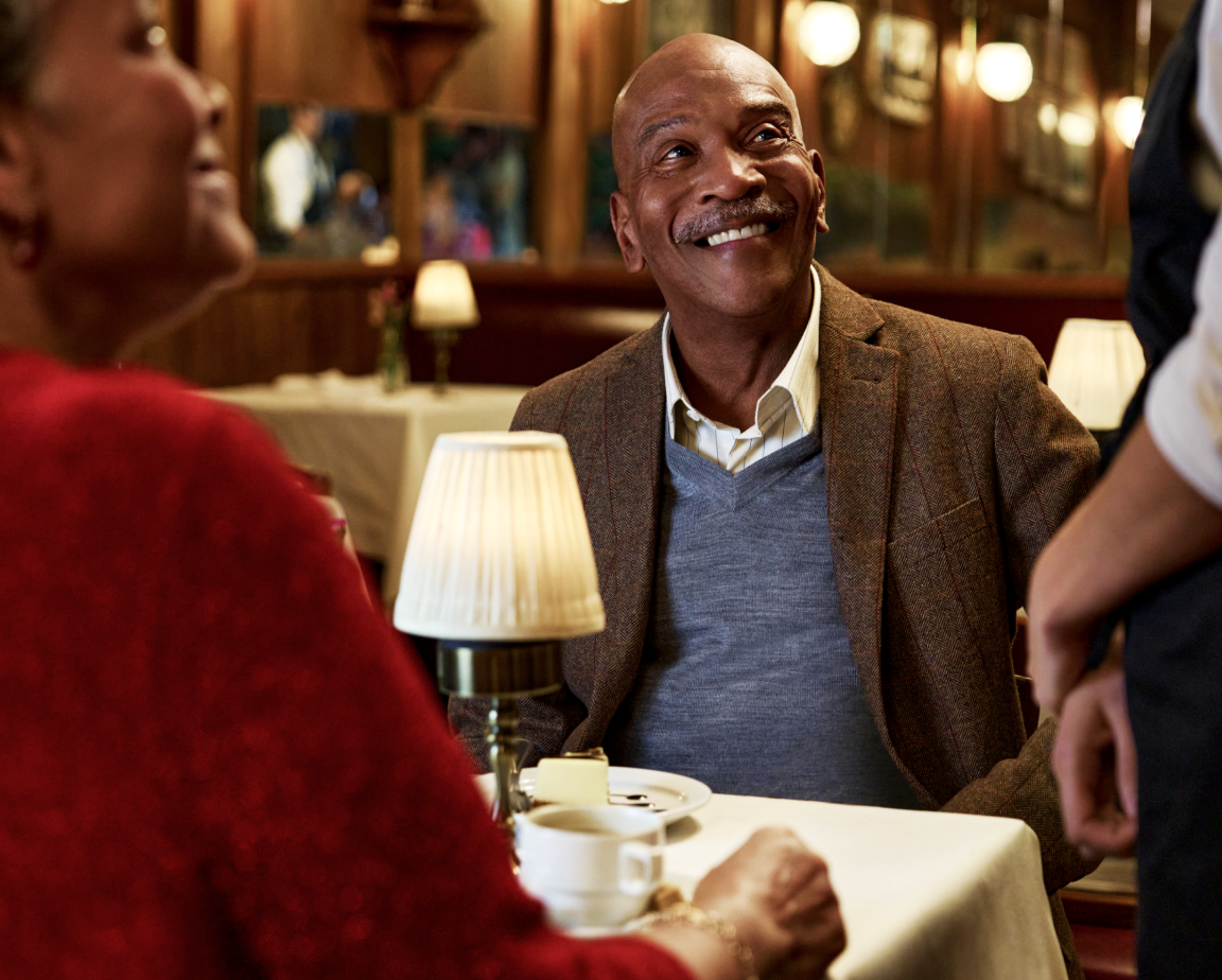 A man smiles while sitting at a table in a dimly lit restaurant, engaging with a waiter. A lit table lamp and a cup of coffee are on the table.