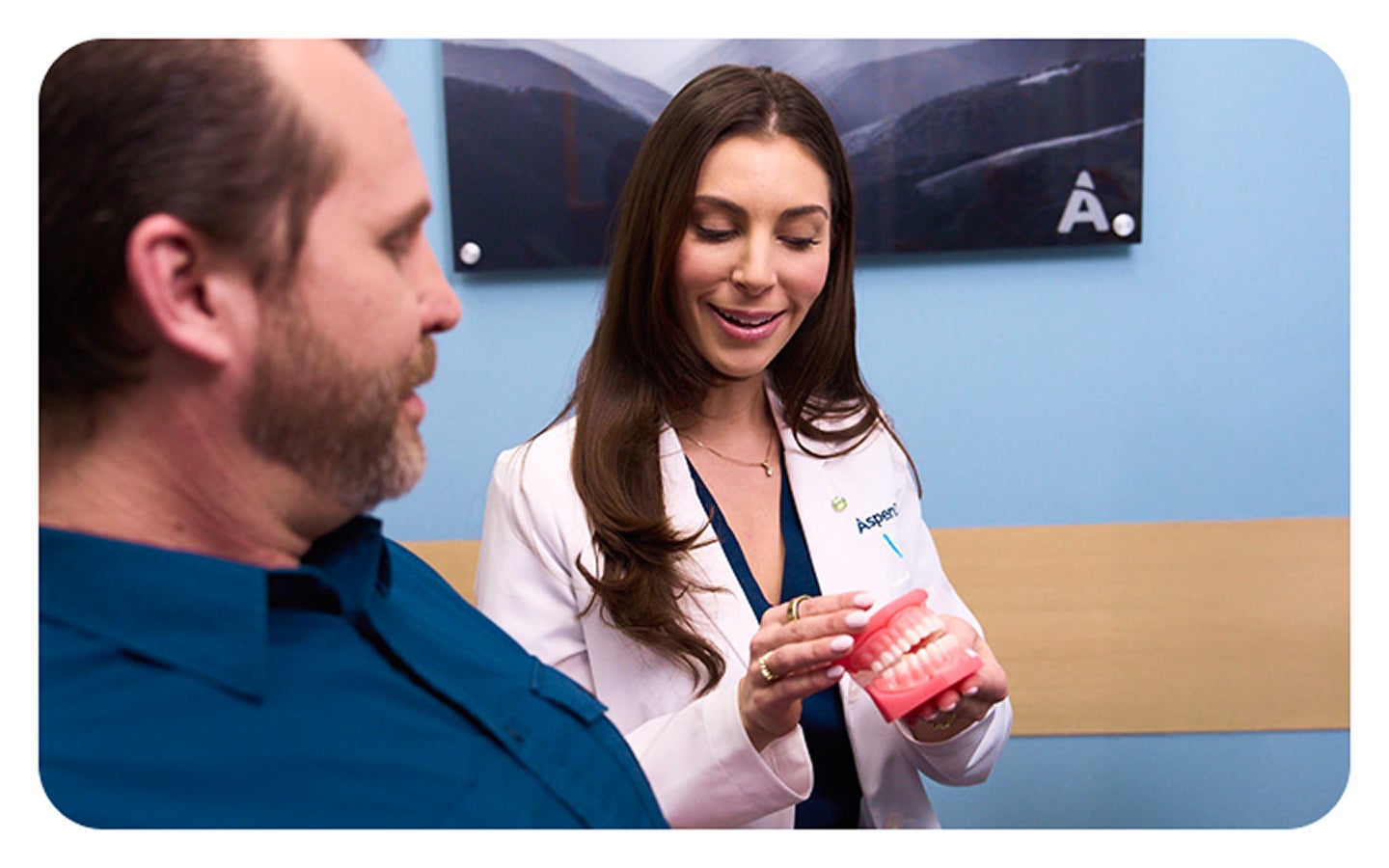 An Aspen Dental dentist in a white coat discusses full arch dentures with a patient, using a model to explain the benefits. The image highlights personalized care and comprehensive tooth replacement options.