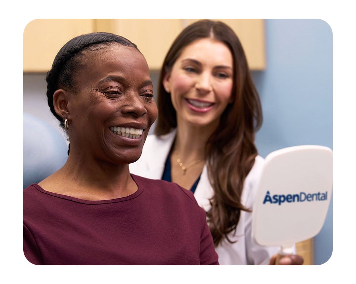 A woman is smiling while looking at her teeth in an Aspen Dental mirror