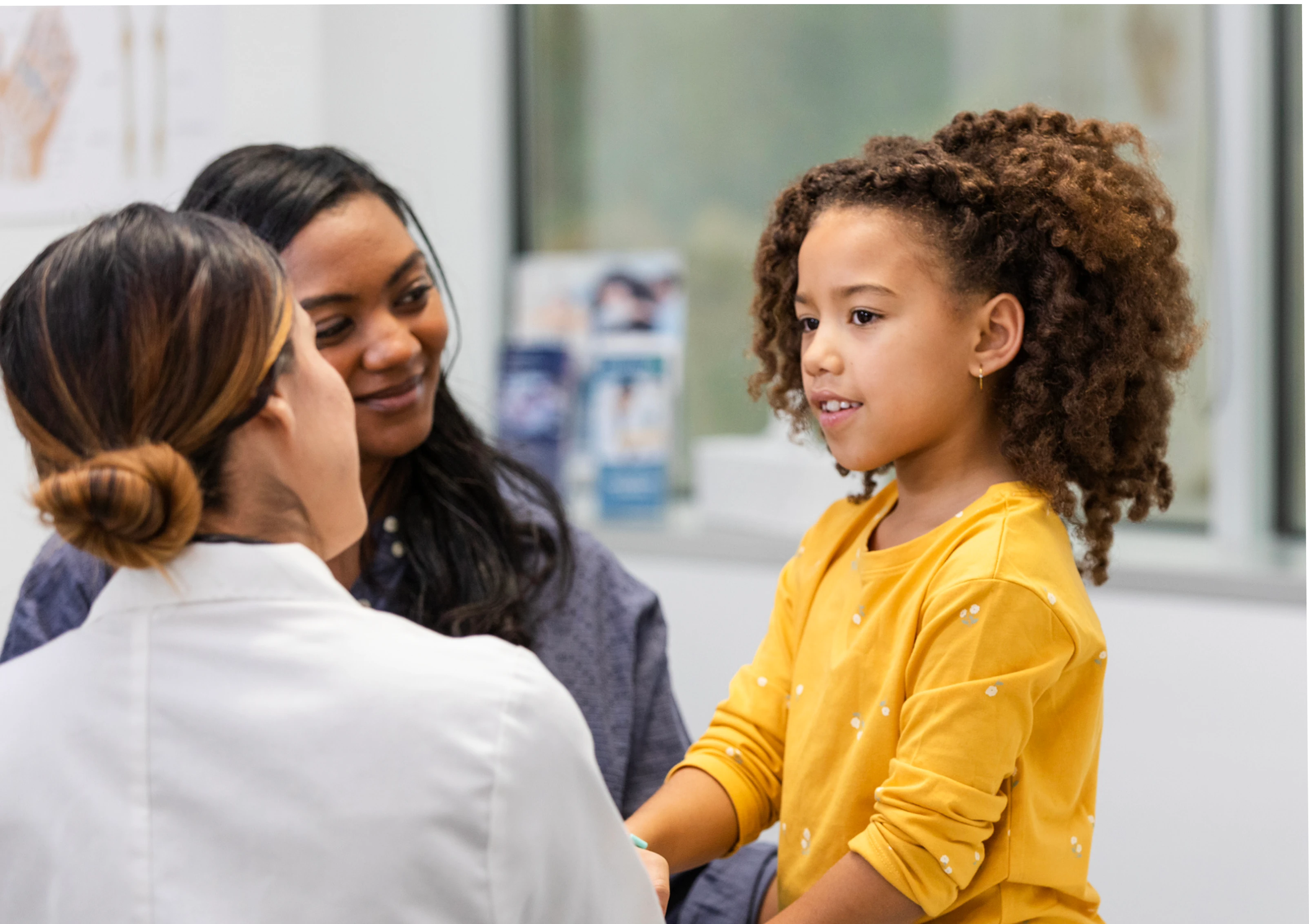 A child sitting in an urgent care clinic looking at a doctor, whose back is to the camera. The parent’s child is looking at the child and smiling.