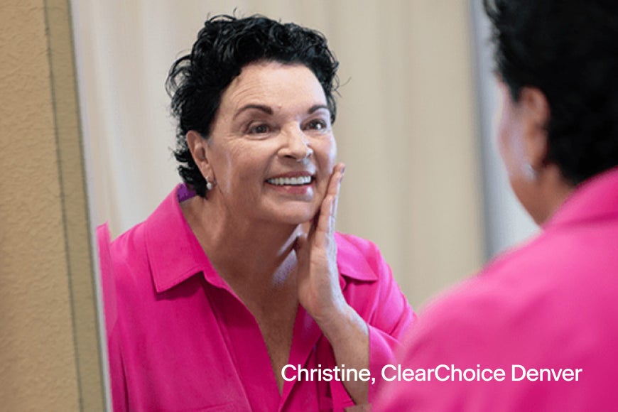 Christine, a ClearChoice Denver patient, smiling and touching her face while looking in the mirror after a successful dental implant procedure.