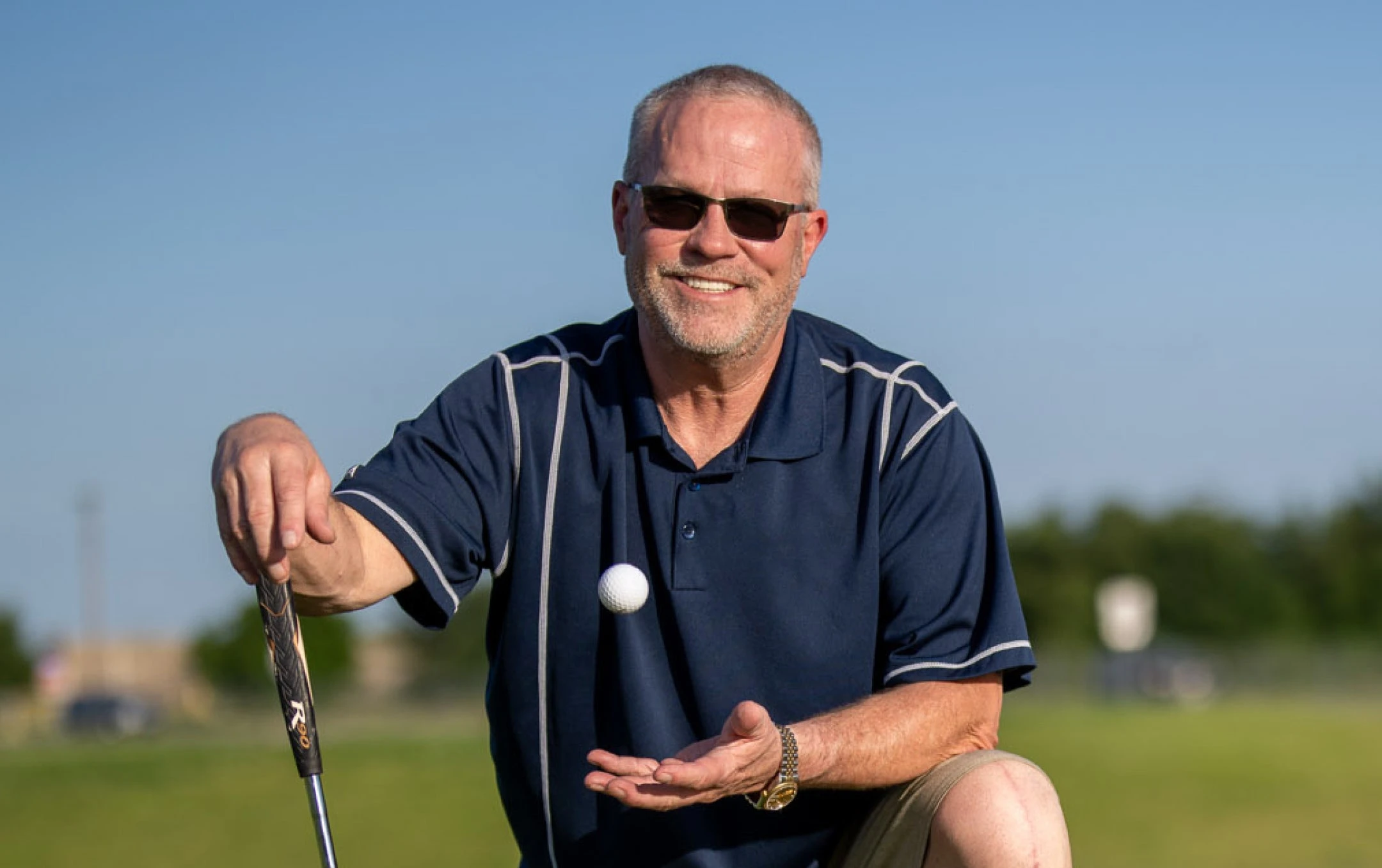 A smiling man wearing sunglasses and a navy blue polo shirt kneeling on a golf course, holding a golf ball and putter under bright sunlight.