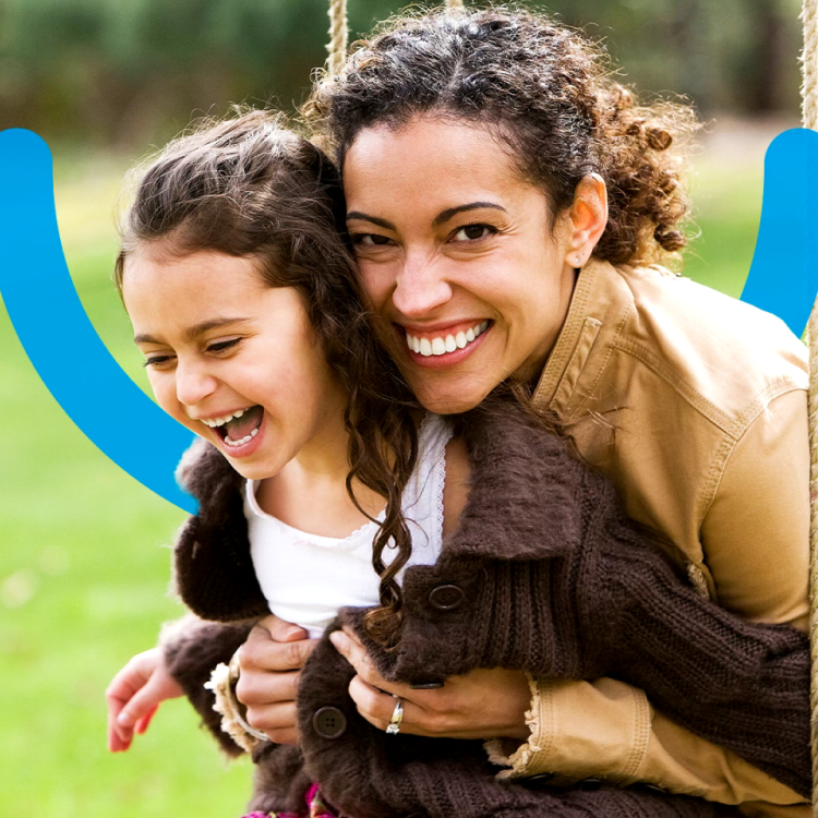 A mother and her little girl sharing a joyful moment on a swing in a park, showcasing a confident smile with an Aspen Dental smile icon behind them.