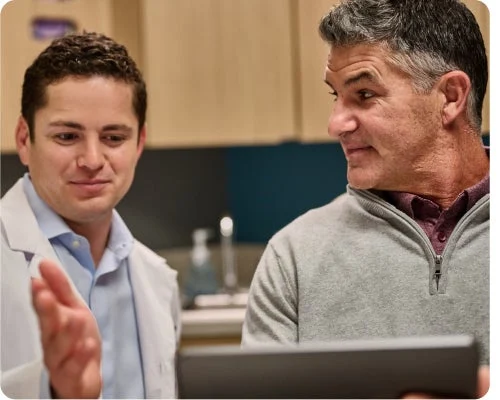 A dentist in a white coat discusses information on a tablet with an older man wearing a grey sweater in a medical office setting.