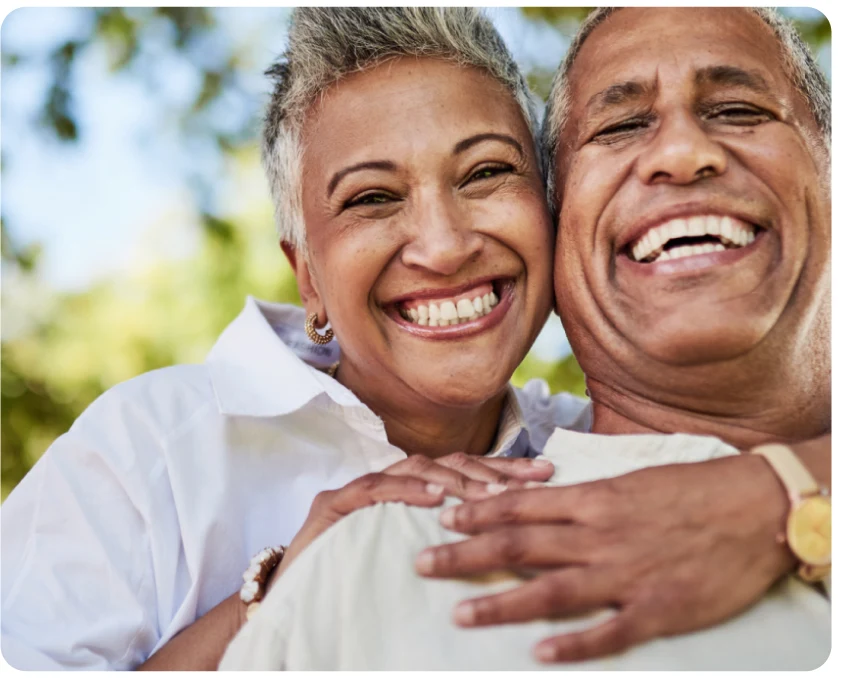 An older couple smiling joyfully, with the woman hugging the man from behind against a clear sky background.