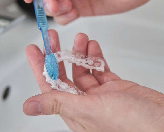 A person holding a clear aligner retainer and cleaning with a toothbrush over the sink.