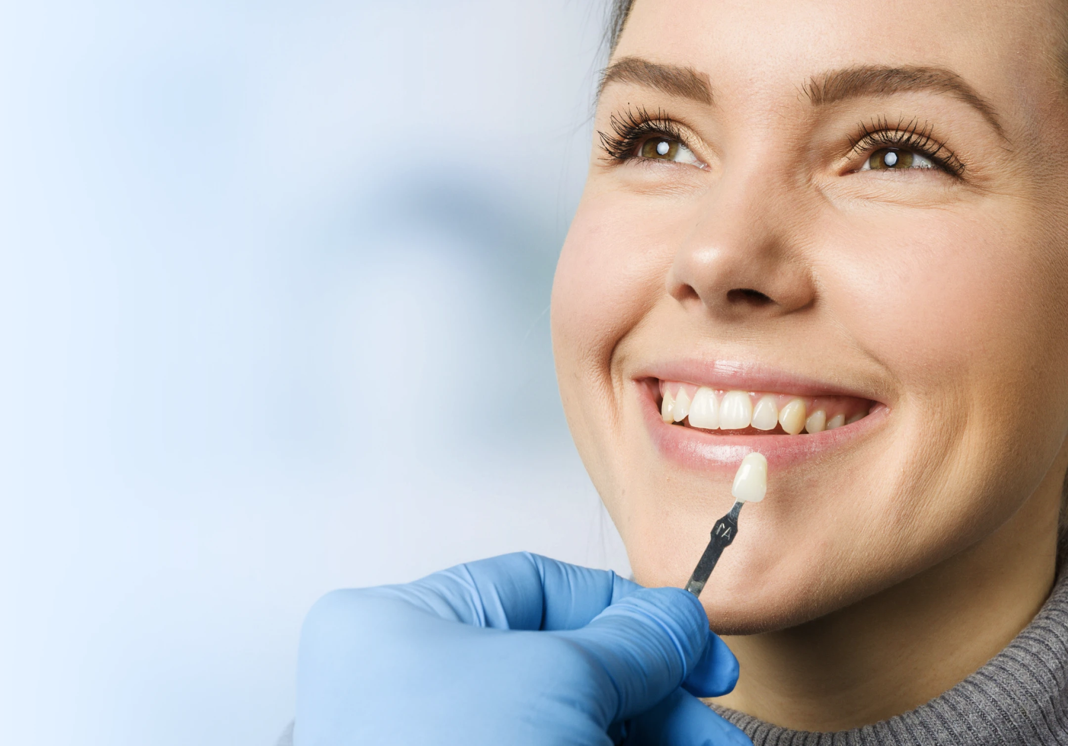 A dental professional wearing blue gloves holds a color chart for veneers against a woman's teeth to ensure precise color matching.