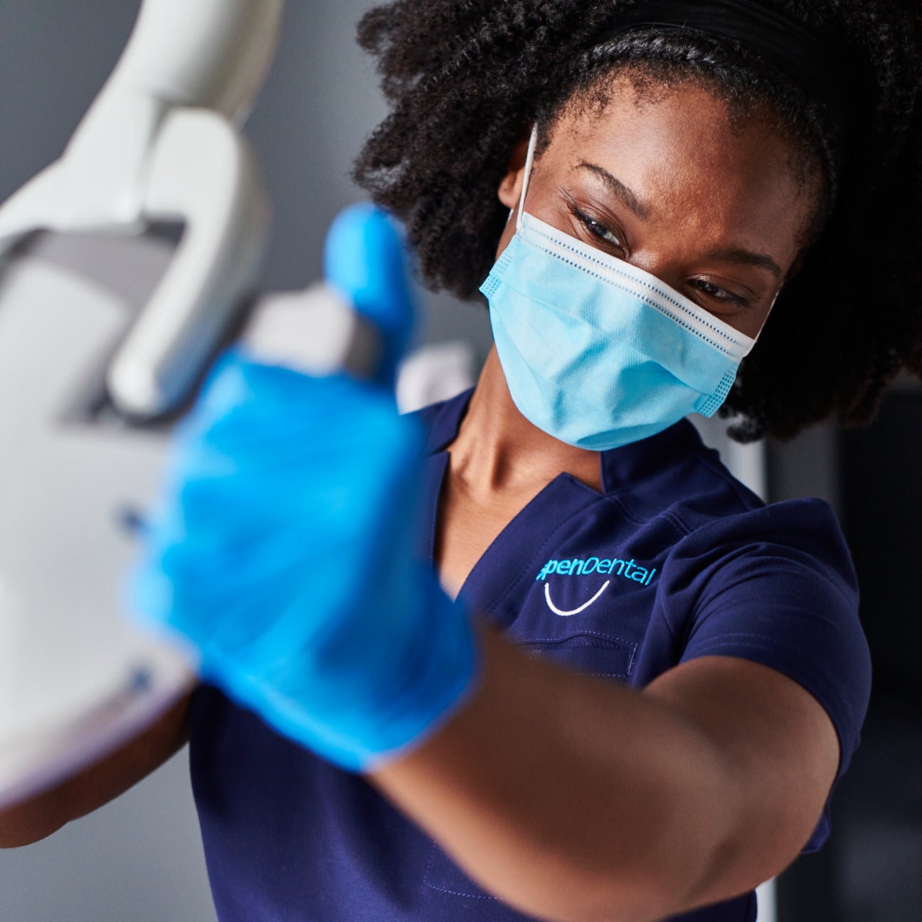 Aspen Dental team member wearing a mask and gloves while adjusting dental equipment, showcasing professionalism and care in a clinical setting.
