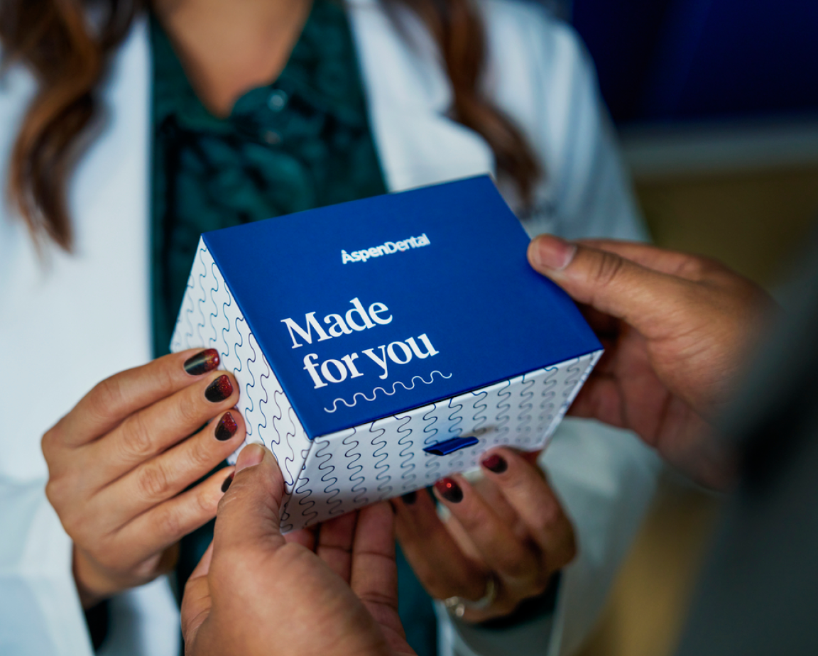 A woman dentist and a man holding a small box labeled "Aspen Dental Made for You." The dentist wears a white coat, suggesting they are in the Aspen Dental office.