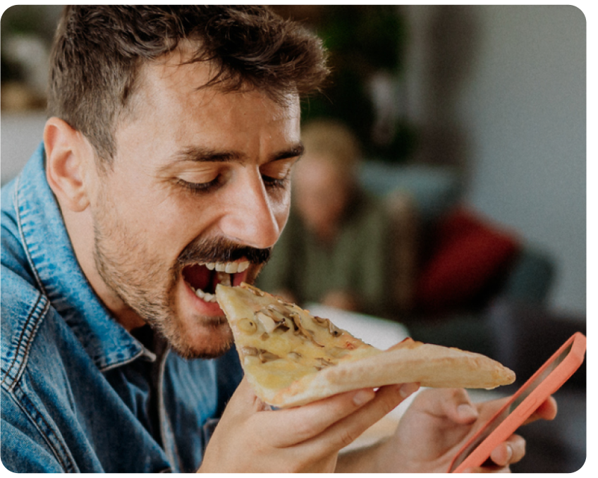 A man wearing a denim shirt eats a slice of pizza while holding a smartphone in his other hand.