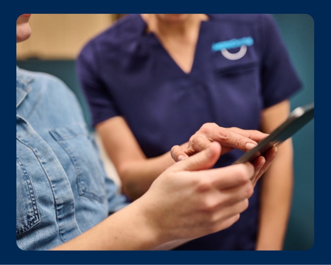 Two people in a healthcare setting look at a handheld device. One person points at the screen while the other holds the device.
