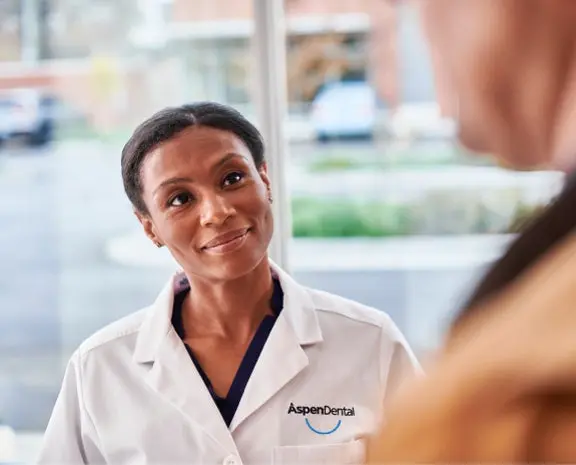 A friendly Aspen Dental professional in a white coat smiling warmly while engaging with a patient, creating a welcoming and reassuring environment at the dental office.