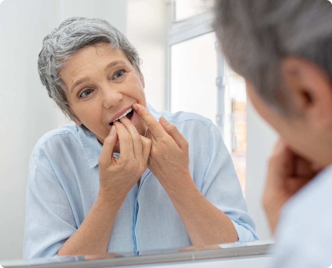 Senior woman standing in front of a mirror carefully flossing her teeth, emphasizing ClearChoice’s recommendation for maintaining gum health and preventing gum disease through regular flossing.