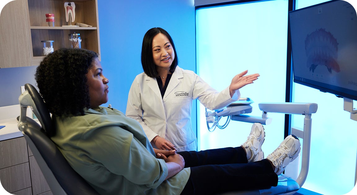 A ClearChoice prosthodontist in a white lab coat explaining to her patient, a woman wearing a green jacket and sitting in a dental chair, her examination results and explaining dental implant procedures and costs.