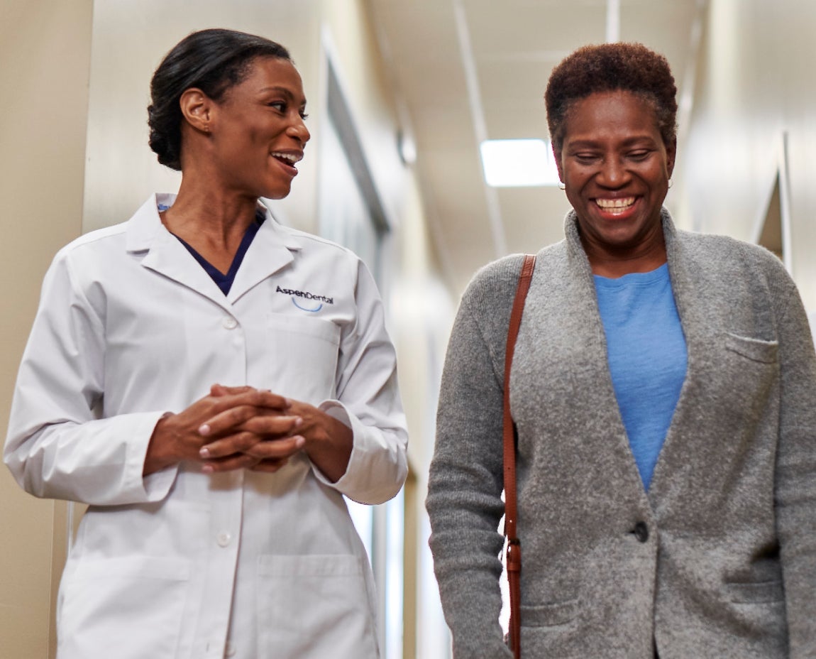 A dentist in an Aspen Dental coat walking and conversing with a smiling patient in a hallway after a dental implant consultation.