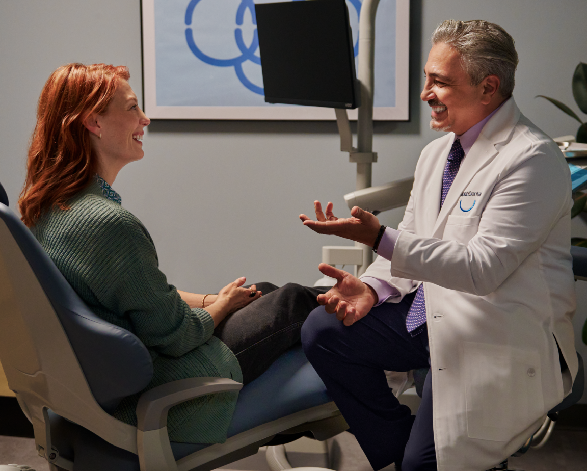 Smiling male dentist in white coat talking with a smiling red-haired female patient in a dental chair.