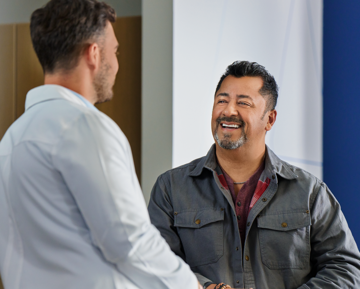 A man with a goatee smiling and speaking with a dentist in a clinical setting.