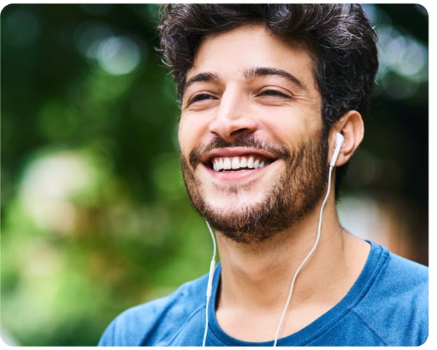 A man in a blue t-shirt smiling with headphones in an outdoor setting.