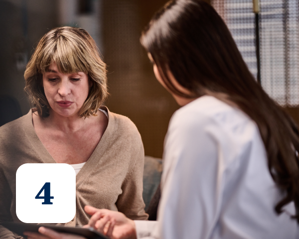 A woman with shoulder-length hair listens as another woman, dressed in a white coat, speaks and points to a tablet. A house icon with the number 4 is in the bottom left corner of the image.