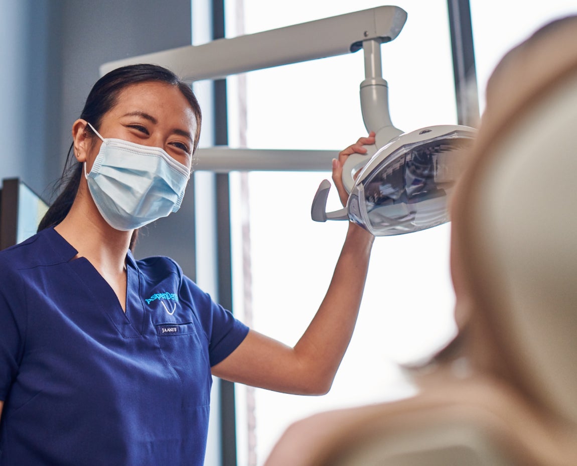 Dentist or dental hygienist wearing a mask and navy scrubs adjusts an overhead light while a patient sits in the dental chair.