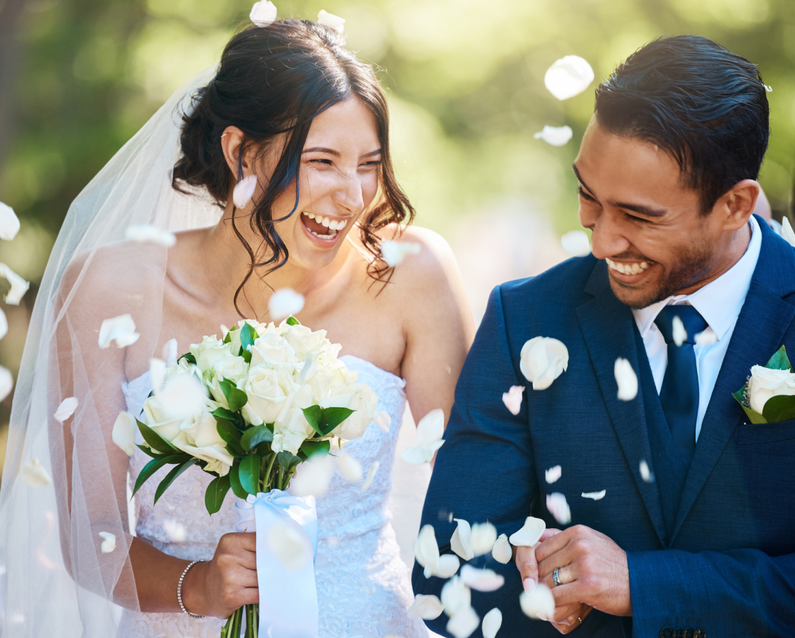 A bride and groom smile joyfully as flower petals are thrown around them. The bride holds a bouquet of white roses and wears a veil; the groom is in a blue suit.