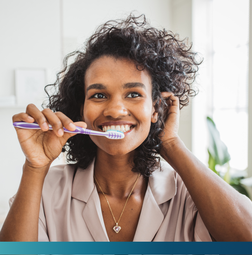 A woman with curly hair smiles while brushing her teeth with a purple toothbrush. She wears a light-colored top and a heart-shaped pendant necklace.