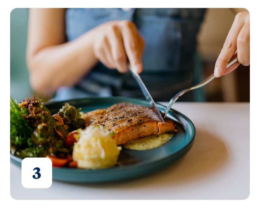 A photo of a woman in a blue sleeveless button-up, cutting into a piece of salmon, on a blue plate, using a fork and knife.
