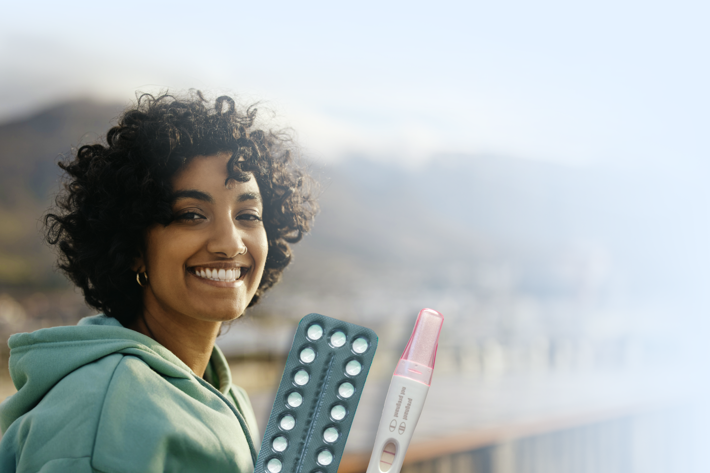 A woman wearing a green hoodie standing outdoors with blurry mountains in the background, and a blister pack of pills and a pregnancy test in the foreground.