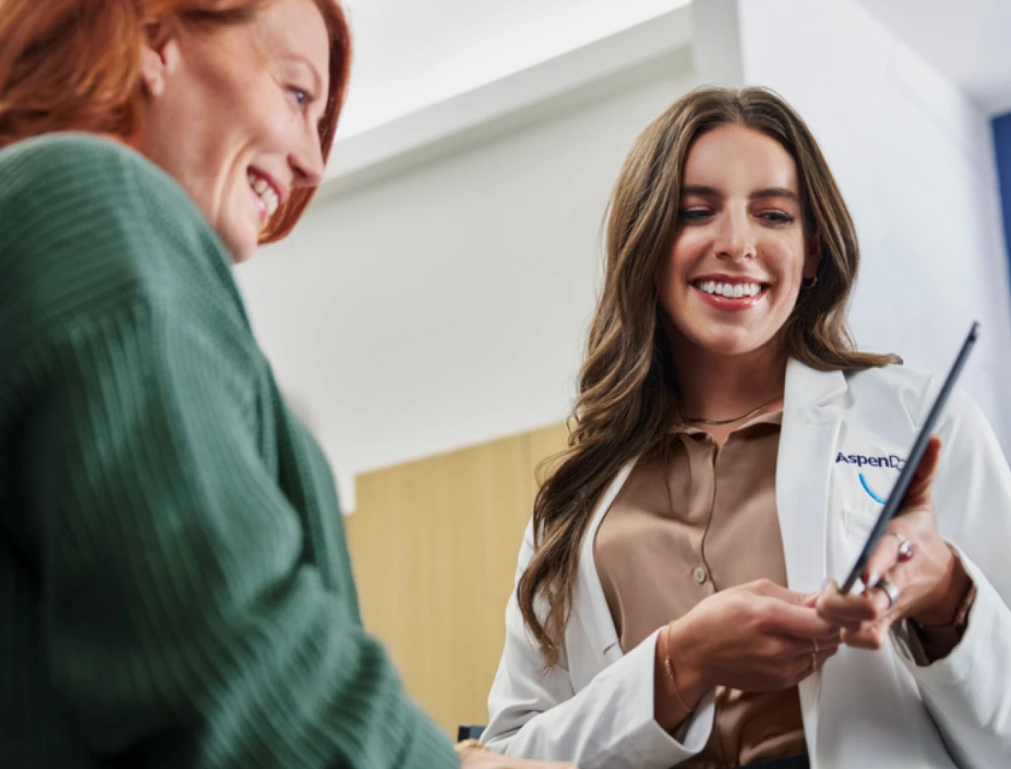 A doctor in a white coat with Aspen Dental logo shows a tablet to a patient with long brown hair, both smiling in a medical office setting.