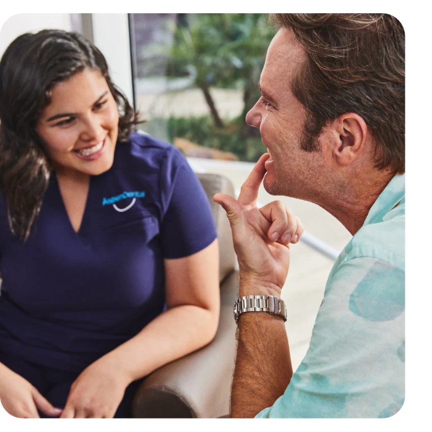 A man and a woman sit in a brightly lit room engaged in conversation. The woman, wearing a Aspen Dental navy scrubs, and listening attentively as the man points towards his teeth, discussing wisdom teeth removal.