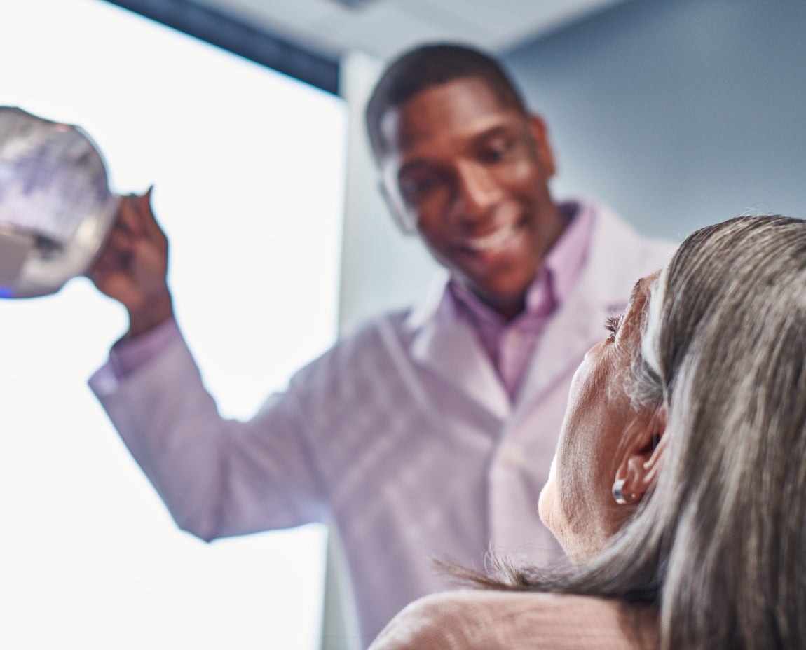 A dentist in a white coat is holding a clipboard and smiling at an older patient during a consultation in a well-lit room.