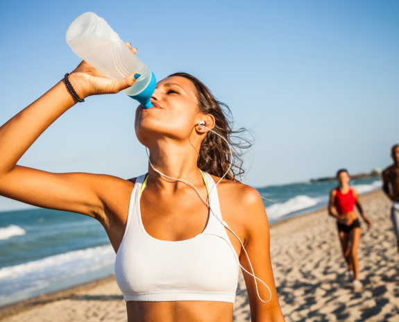 Woman drinking water while running on the beach, wearing workout attire and earbuds under a clear blue sky.