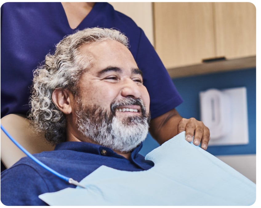 A man with curly gray hair and a beard sits in a dental chair, smiling, while a dental professional stands next to him with a hand on his shoulder.