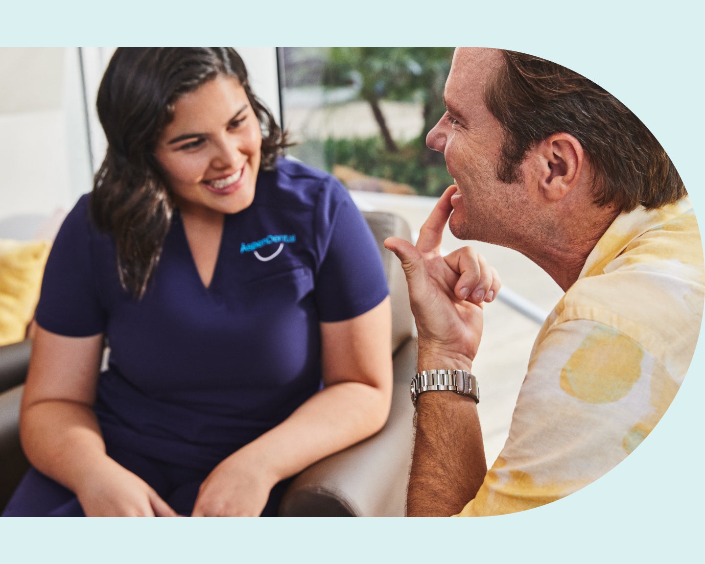  man and a woman in scrubs sit in a dental office. The man points to his teeth while talking to the woman.