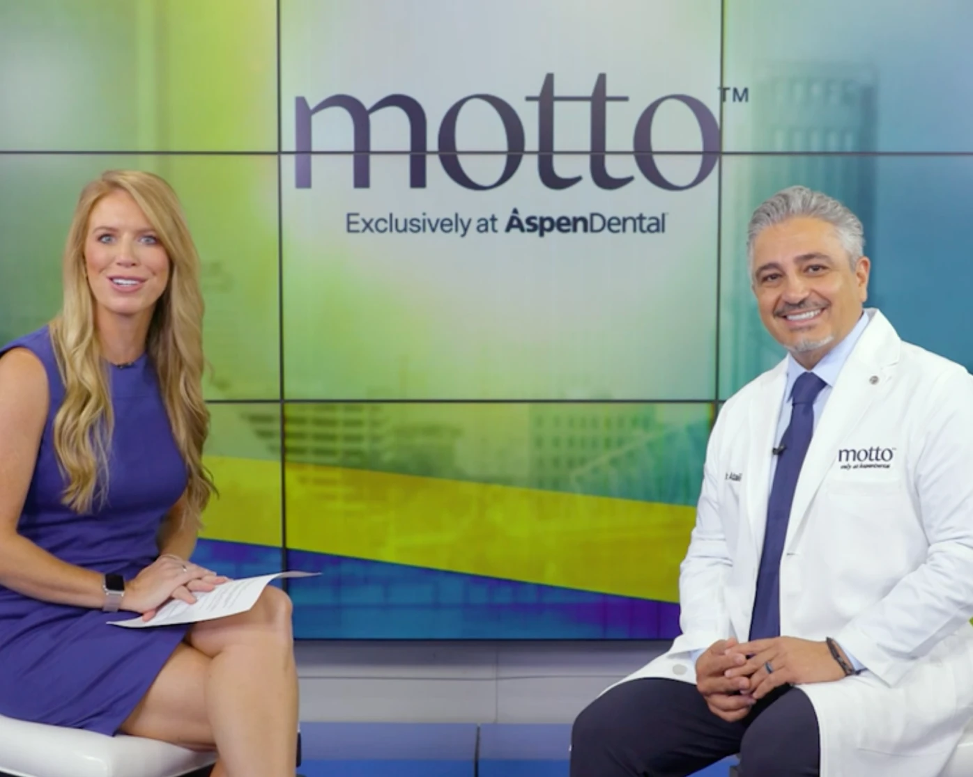 Dr. Ataii sits next to a smiling news anchor, with the text "Motto: Exclusively at Aspen Dental" in the background