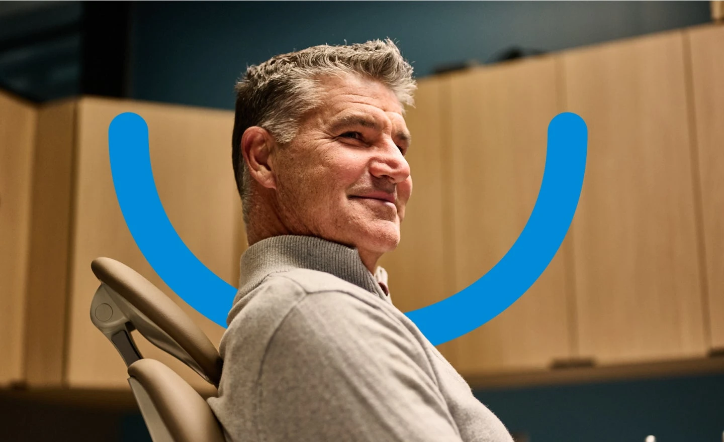 A man with gray hair is seated in an office chair, smiling slightly. There is a blue curved graphic behind him on the wall depicting a smile.