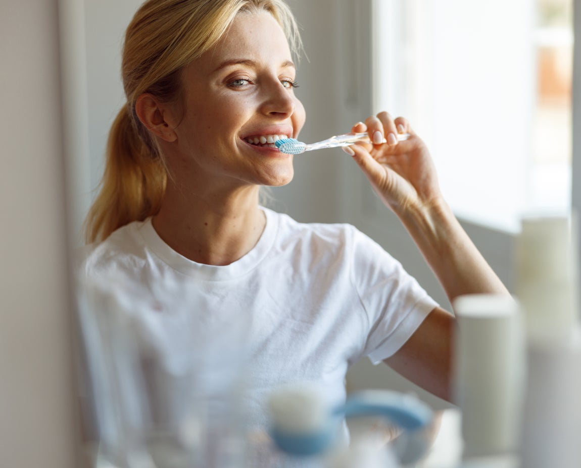 A woman wearing a white t-shirt is brushing her teeth while looking in a mirror.